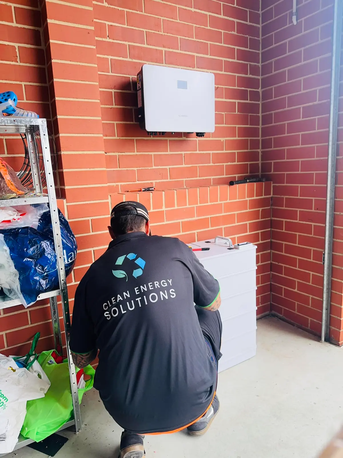 A CES electrician crouched beside a home battery, wiring it under a wall-mounted inverter on a red brick wall