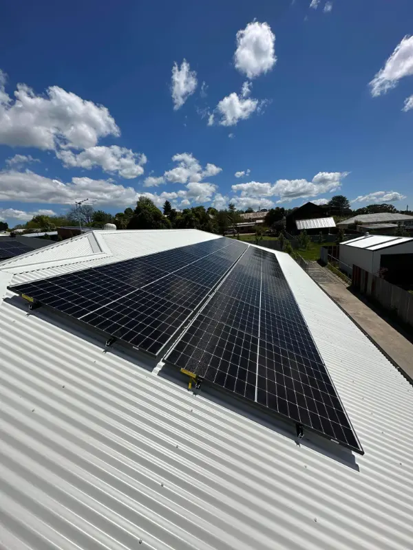 Two rows of solar panels on a white corrugated roof under a bright blue sky with clouds