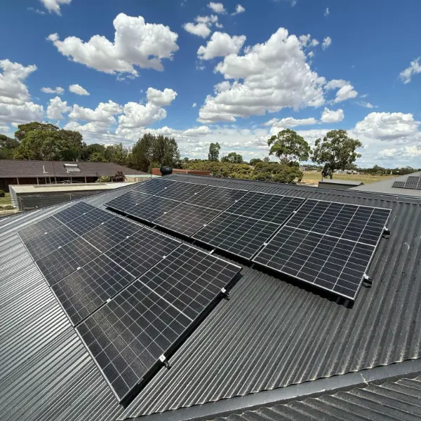 Solar panels on a dark corrugated roof with gum trees and a cloudy blue sky behind